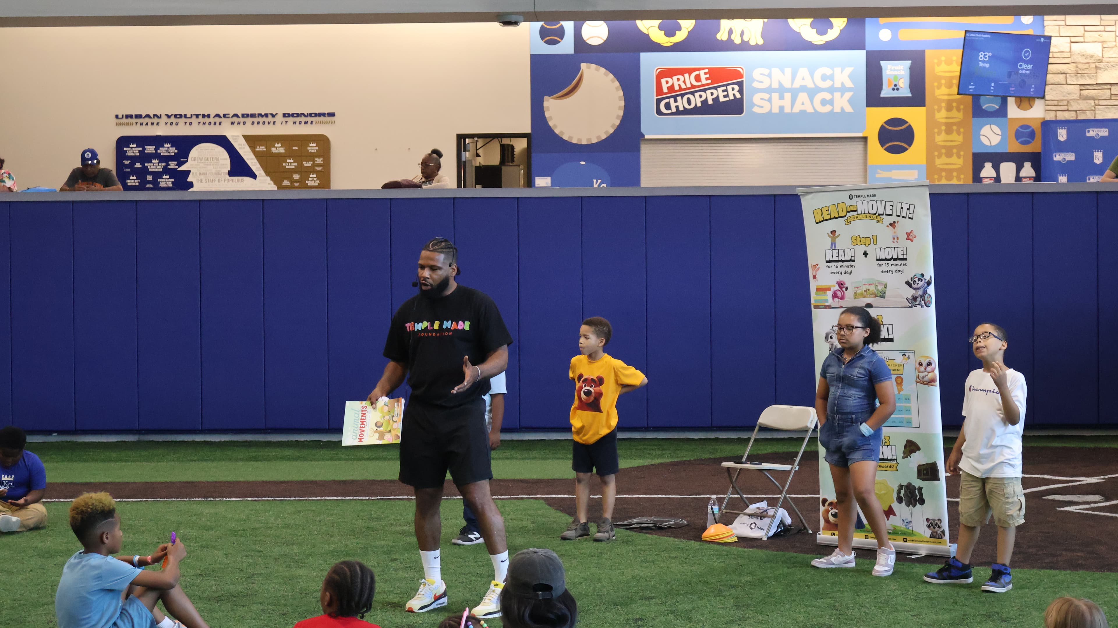 Tony Temple holding a book and leading Read and Move It with children on the indoor field at the Kansas City Urban Youth Academy